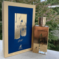 Maison Alhambra Minerale Gold by Maison Alhambra 100ml EDP — A gold-colored perfume bottle and its matching yellow and blue box are displayed side-by-side against a white background. | Maison Alhambra Minerale Gold by Maison Alhambra 100ml EDP — Botol minyak wangi berwarna emas dan kotak padanannya yang berwarna