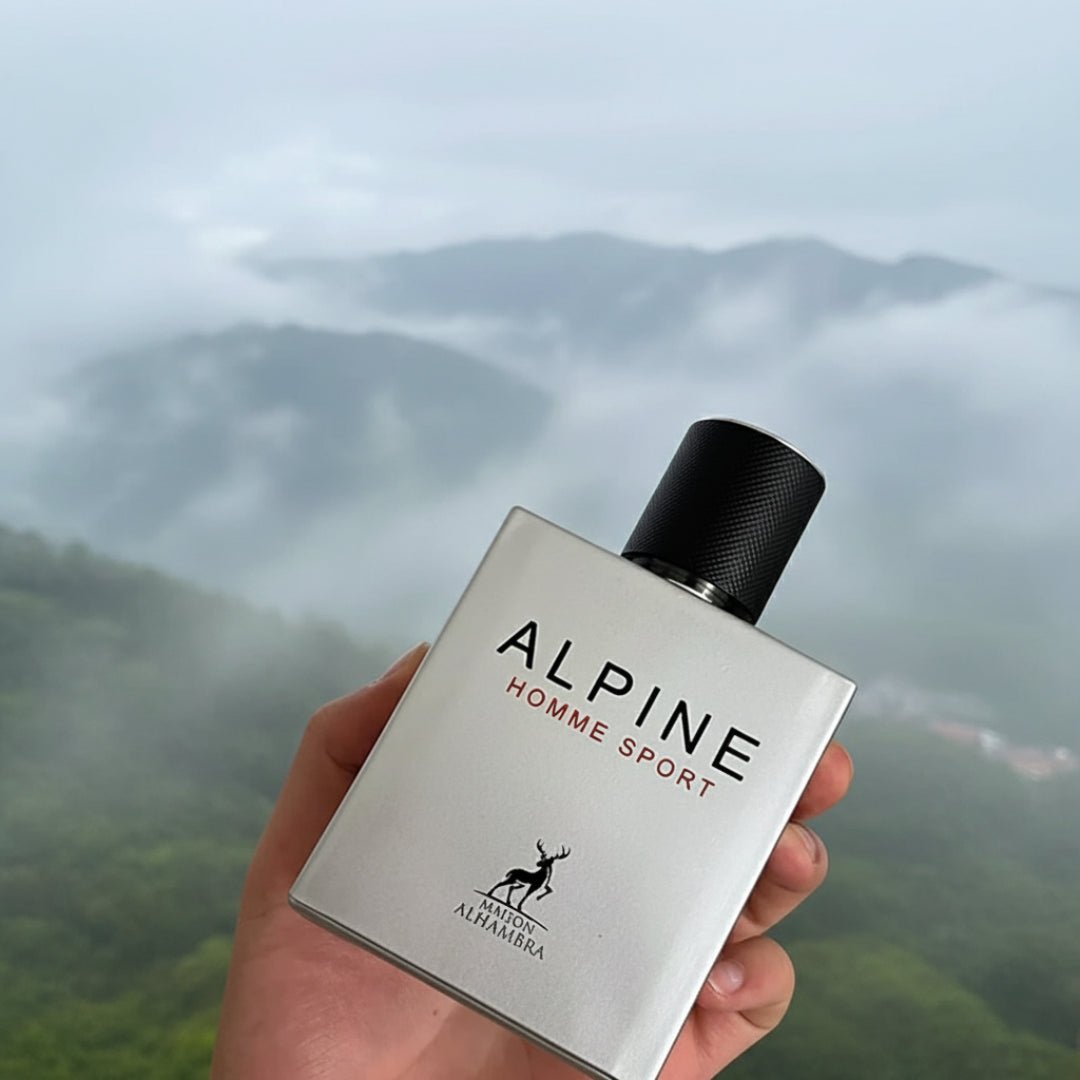 Maison Alhambra Alpine Homme Sport By Maison Alhambra Men 100ml — A hand holds a silver perfume bottle with a black cap against a misty mountain landscape. | Maison Alhambra Alpine Homme Sport By Maison Alhambra Men 100ml — Tangan memegang botol minyak wangi perak dengan penutup hitam berlatarkan landskap pergunungan