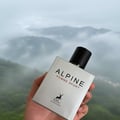 Maison Alhambra Alpine Homme Sport By Maison Alhambra Men 100ml — A hand holds a silver perfume bottle with a black cap against a misty mountain landscape. | Maison Alhambra Alpine Homme Sport By Maison Alhambra Men 100ml — Tangan memegang botol minyak wangi perak dengan penutup hitam berlatarkan landskap pergunungan