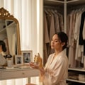 Maison Alhambra Anarch by Maison Alhambra 100ml EDP Women — Woman in a silk robe smelling a perfume bottle with a gold tassel, sitting at a vanity with a mirror and closet in the background. | Maison Alhambra Anarch by Maison Alhambra 100ml EDP Women — Wanita berbaju sutera menghidu botol minyak wangi bertali emas