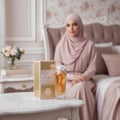 Maison Alhambra Athena Eau de Parfum 100 ml Ville Femme — A bottle of golden perfume and its marble-patterned box sit on a white marble table, with a woman in a pink hijab blurred in the background. | Maison Alhambra Athena Eau de Parfum 100 ml Ville Femme — Sebotol minyak wangi keemasan dan kotaknya yang bermotif