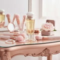 Maison Alhambra Athena Eau de Parfum 100 ml Ville Femme — A perfume bottle sits on a rose gold vanity table surrounded by makeup brushes, a compact mirror, a pearl bracelet, and a ring, with a pink peony in a jewelry box in the background. | Maison Alhambra Athena Eau de Parfum 100 ml Ville Femme — Sebotol minyak