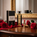 Maison Alhambra B.A.D. Femme — A gold and black perfume bottle and its box sit on a wooden dresser, surrounded by red velvet fabric and red roses, with a lipstick and a mirror in the background. | Maison Alhambra B.A.D. Femme — Botol dan kotak minyak wangi emas dan hitam terletak di atas meja kayu, dikelilingi fabrik