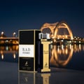 Maison Alhambra B.A.D. Femme — A dark blue and gold perfume bottle and its box stand on a reflective surface at night, with a brightly lit bridge and water in the background. | Maison Alhambra B.A.D. Femme — Botol dan kotak minyak wangi biru gelap dan emas berdiri di permukaan reflektif pada waktu malam, dengan