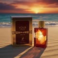 Maison Alhambra Tobacco Your Touch – A golden box and a glass perfume bottle with amber liquid sit on a sandy beach at sunset with the ocean in the background. | Maison Alhambra Tobacco Your Touch – Kotak emas dan botol minyak wangi kaca dengan cecair ambar diletakkan di pantai berpasir semasa matahari terbenam dengan