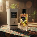 Maison Alhambra The Tux — A white perfume box and a clear glass bottle of The Tux Eau de Parfum are displayed on a patterned fabric surface, with sparkling effects around the bottle. | Maison Alhambra The Tux — Peti kotak minyak wangi putih dan sebotol kaca jernih The Tux Eau de Parfum dipamerkan di atas permukaan