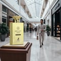 Maison Alhambra Brulant Oud for Men 100ml — A tall, golden perfume bottle stands on a wooden pedestal in a modern shopping mall, with a man in a light suit walking in the background. | Maison Alhambra Brulant Oud untuk Lelaki 100ml — Botol minyak wangi emas tinggi berdiri di atas alas kayu di pusat membeli-belah