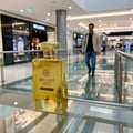 Maison Alhambra Brulant Oud — A golden perfume bottle with a decorative cap sits on a reflective surface in a modern shopping mall with a man walking in the background. | Maison Alhambra Brulant Oud — Botol minyak wangi keemasan dengan penutup hiasan terletak di permukaan reflektif di pusat membeli-belah moden dengan