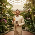 Maison Alhambra Brulant Oud Maison Alhambra for Men 100ml — Man in a cream suit holding a gold perfume bottle and its box in a lush indoor garden with orchids and a stream. | Maison Alhambra Brulant Oud Maison Alhambra for Men 100ml — Lelaki berbaju krim memegang botol minyak wangi emas dan kotaknya di taman dalaman