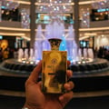 Maison Alhambra Brulant Oud — A hand holds a golden perfume bottle in front of a blurred mall fountain and lights. | Tangan memegang botol minyak wangi emas di hadapan air pancut pusat membeli-belah yang kabur dan lampu.