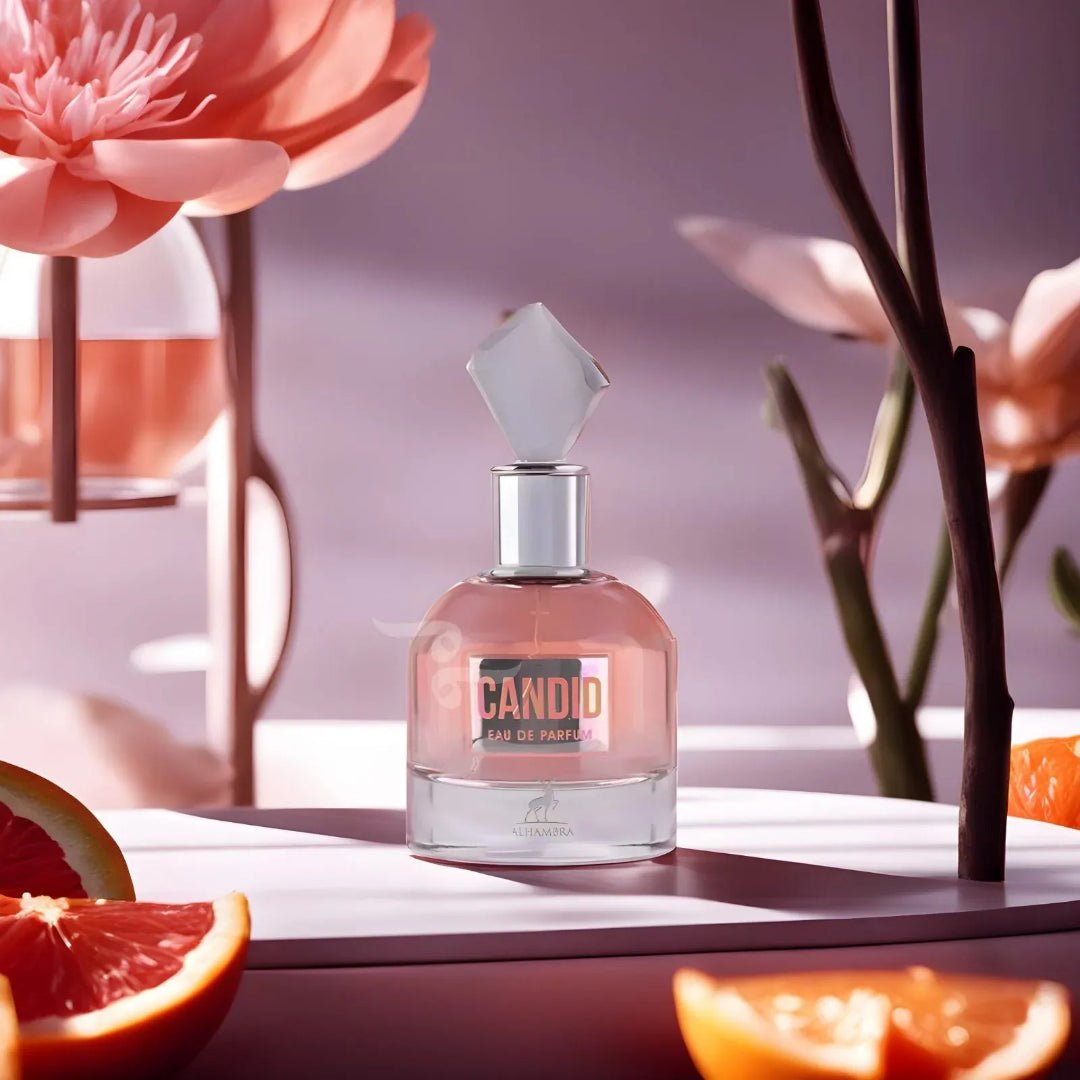 Maison Alhambra Candid by Maison Alhambra 100 ml for Women — A pink perfume bottle with a faceted clear cap sits on a white surface, surrounded by slices of grapefruit and orange, with blurred pink flowers and branches in the background. | Maison Alhambra Candid by Maison Alhambra 100 ml for Women — Botol minyak wangi