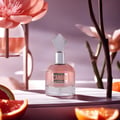Maison Alhambra Candid by Maison Alhambra 100 ml for Women — A pink perfume bottle with a faceted clear cap sits on a white surface, surrounded by slices of grapefruit and orange, with blurred pink flowers and branches in the background. | Maison Alhambra Candid by Maison Alhambra 100 ml for Women — Botol minyak wangi