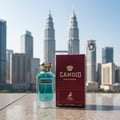 Maison Alhambra Candid Pour Homme — A clear glass perfume bottle with blue liquid and a gold cap stands next to its maroon box, with the Petronas Twin Towers and city skyline in the background. | Maison Alhambra Candid Pour Homme — Botol minyak wangi kaca jernih dengan cecair biru dan penutup emas berdiri di sebelah
