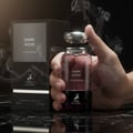 Maison Alhambra Dark Aoud — Hand holding a glass perfume bottle with a silver cap, surrounded by smoke effects, with the product box in the background. | Tangan memegang botol minyak wangi kaca bertutup perak, dikelilingi kesan asap, dengan kotak produk di latar belakang.