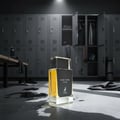 Maison Alhambra Dark Door Intense by Maison Alhambra 100ml Men — A clear glass perfume bottle with yellow liquid sits on a wet concrete floor in a locker room, with lockers and gym equipment in the background. | Maison Alhambra Dark Door Intense by Maison Alhambra 100ml Men — Botol minyak wangi kaca jernih berisi