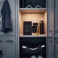 Maison Alhambra Dark Door Intense — A dark perfume bottle and its box sit on a shelf in a locker, next to a dark water bottle. A dark blue towel hangs on the left locker door. | Maison Alhambra Dark Door Intense — Botol minyak wangi gelap dan kotaknya diletakkan di rak dalam loker, bersebelahan dengan botol air gelap.