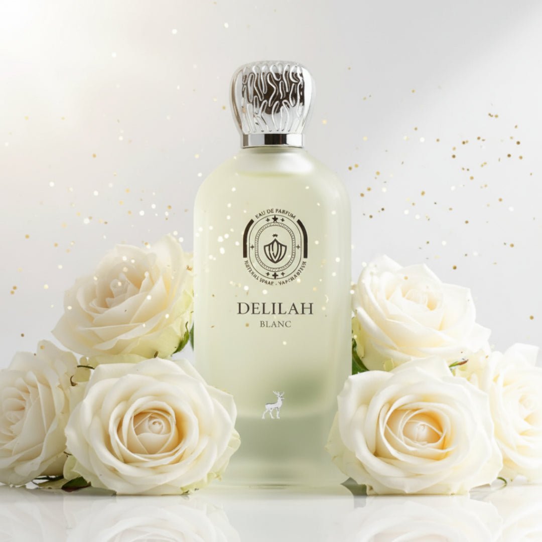 Maison Alhambra Delilah Blanc by Maison Alhambra 100ml EDP — A frosted glass perfume bottle with a silver ornate cap, surrounded by white roses and gold glitter against a white background. | Maison Alhambra Delilah Blanc by Maison Alhambra 100ml EDP — Botol kaca beku dengan penutup perak berhias, dikelilingi mawar