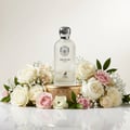 Maison Alhambra Delilah Blanc by Maison Alhambra 100ml EDP — A frosted glass perfume bottle with a silver cap sits on a gold pedestal, surrounded by a lush arrangement of white and pink roses and baby's breath. | Maison Alhambra Delilah Blanc by Maison Alhambra 100ml EDP — Botol kaca beku dengan penutup perak terletak