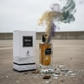 Ebene by Maison Alhambra Perfume 90 ml EDP — A cracked glass perfume bottle with colored smoke emanating from it, standing next to its white box on a concrete surface with shattered glass fragments. | Ebene by Maison Alhambra Perfume 90 ml EDP — Botol kaca pecah dengan asap berwarna keluar daripadanya, berdiri di