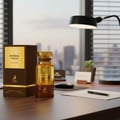 Maison Alhambra Eternal Touch — A golden perfume bottle and its brown box sit on a wooden desk with a black lamp and a pen on a notepad, overlooking a city skyline at dusk. | Maison Alhambra Eternal Touch — Botol minyak wangi keemasan dan kotaknya yang berwarna coklat diletakkan di atas meja kayu dengan lampu hitam