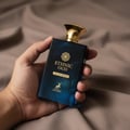 Maison Alhambra Ethnic Oud by Maison Alhambra 100ml EDP — A hand holds a dark blue glass perfume bottle with a gold cap against a textured brown fabric background. | Maison Alhambra Ethnic Oud by Maison Alhambra 100ml EDP — Tangan memegang botol minyak wangi kaca biru gelap dengan penutup emas berlatarkan fabrik