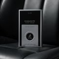 Maison Alhambra Exquisite Club By Maison Alhambra For Men 100ml — A black and silver perfume box with the text "EXQUISITE POUR HOMME" and a deer logo is placed on a dark, textured surface, possibly leather. | Maison Alhambra Exquisite Club By Maison Alhambra For Men 100ml — Kotak minyak wangi hitam dan perak dengan