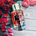 Maison Alhambra Exclusif Rose Maison Alhambra 100ml EDP — A clear glass perfume bottle with a gold cap and a dark red label sits on a wooden surface surrounded by red rose petals and leaves. | Maison Alhambra Exclusif Rose Maison Alhambra 100ml EDP — Botol minyak wangi kaca jernih bertudung emas dan berlabel merah