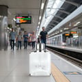 Maison Alhambra Expose Blanc by Maison Alhambra 100ml EDP Men — A white perfume bottle sits on a train station platform with people walking in the background. | Maison Alhambra Expose Blanc by Maison Alhambra 100ml EDP Men — Botol minyak wangi putih diletakkan di platform stesen kereta api dengan orang ramai berjalan