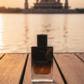 Maison Alhambra Exquisite Prive 100 ml EDP Men — A black and clear glass perfume bottle with gold accents sits on a wooden deck with a blurred background of water and a building at sunset. | Maison Alhambra Exquisite Prive 100 ml EDP Men — Botol kaca hitam dan jernih dengan aksen emas diletakkan di atas dek kayu