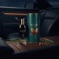 Maison Alhambra Glacier Bold by Maison Alhambra 100ml EDP — A dark glass perfume bottle and its teal cylindrical case sit on a wooden surface inside a car, with green sprigs and wood shavings. | Maison Alhambra Glacier Bold by Maison Alhambra 100ml EDP — Botol minyak wangi kaca gelap dan bekas silindernya yang