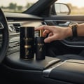 Maison Alhambra Glacier Le Noir — A hand holds a black perfume bottle with a gold cap next to its cylindrical packaging inside a car's center console. | Maison Alhambra Glacier Le Noir — Tangan memegang botol minyak wangi hitam bertutup emas di sebelah pembungkus silindernya di dalam konsol tengah kereta.