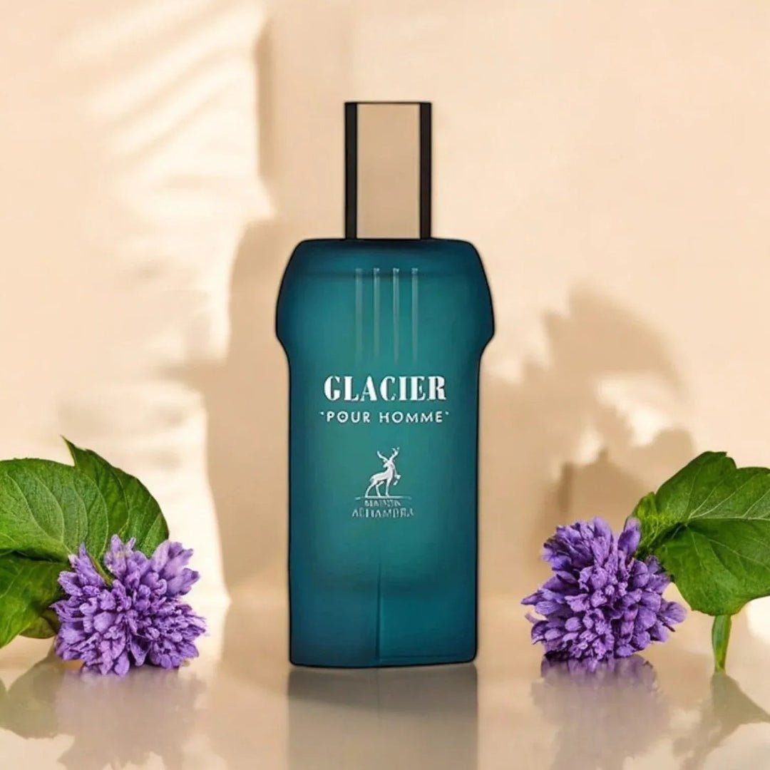 Maison Alhambra Glacier Pour Homme — A teal perfume bottle with a gold cap stands centered, flanked by purple flowers and green leaves on a reflective surface, with soft shadows in the background. | Maison Alhambra Glacier Pour Homme — Botol minyak wangi teal dengan penutup emas berdiri di tengah, diapit oleh bunga