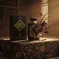 Maison Alhambra Infini Oud Joyous — A black perfume bottle and its box are displayed on a patterned cloth, with oud wood pieces exploding from the bottle. | Maison Alhambra Infini Oud Joyous — Sebotol minyak wangi hitam dan kotaknya dipamerkan di atas kain bermotif, dengan ketulan kayu oud meletup dari botol.