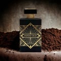 Maison Alhambra Infini Oud by Maison Alhambra Man 100ml EDP — A black perfume bottle with gold geometric art deco detailing and gold textured accents on the cap, placed on a surface with piles of dark brown powder on either side. | Maison Alhambra Infini Oud by Maison Alhambra Man 100ml EDP — Botol minyak wangi hitam