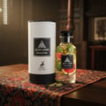 Maison Alhambra Jean Lowe Immortel — A cylindrical white box and a clear glass perfume bottle filled with liquid and citrus slices, placed on a patterned fabric runner on a wooden table. | Maison Alhambra Jean Lowe Immortel — Kotak silinder putih dan botol minyak wangi kaca jernih berisi cecair dan hirisan sitrus