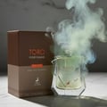 Maison Alhambra Toro Pour Homme Glace – A glass perfume bottle with a light green liquid, surrounded by wisps of white and blue smoke, sits next to its brown box on a marble surface. | Maison Alhambra Toro Pour Homme Glace – Botol minyak wangi kaca berisi cecair hijau muda, dikelilingi kepulan asap putih dan biru