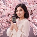 Maison Alhambra Infini Rose by Maison Alhambra 100ml EDP Unisex — A young woman with short dark hair holds a purple perfume bottle with gold geometric designs in front of a pink cherry blossom tree. | Maison Alhambra Infini Rose by Maison Alhambra 100ml EDP Unisex — Seorang wanita muda berambut gelap pendek memegang