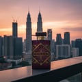 Maison Alhambra Infini Rose — A rectangular burgundy perfume bottle with gold geometric accents and the text "INFINI ROSE" is placed on a railing with a blurred city skyline and sunset in the background. | Botol minyak wangi segi empat berwarna merah anggur dengan perincian geometri emas dan teks "INFINI ROSE"