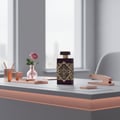 Maison Alhambra Infini Rose by Maison Alhambra 100ml EDP Unisex — A dark purple perfume bottle with gold geometric accents sits on a desk next to a small pink vase with a single pink rose, a rose gold stapler, and rose gold desk organizers. | Maison Alhambra Infini Rose by Maison Alhambra 100ml EDP Unisex — Botol