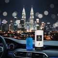Maison Alhambra Jean Lowe Azure by Maison Alhambra EDP 100 ml — Blue perfume bottle and its cylindrical packaging on a car dashboard at night with a city skyline and Petronas Twin Towers in the background. | Maison Alhambra Jean Lowe Azure by Maison Alhambra EDP 100 ml — Botol minyak wangi biru dan pembungkus
