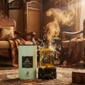 Maison Alhambra Jean Lowe Fraiche — A perfume bottle shattering with golden smoke and glass shards erupting around it, placed on a dark base next to its light green cylindrical packaging, set against a vintage interior with ornate chairs and patterned fabric. | Maison Alhambra Jean Lowe Fraiche — Botol minyak wangi