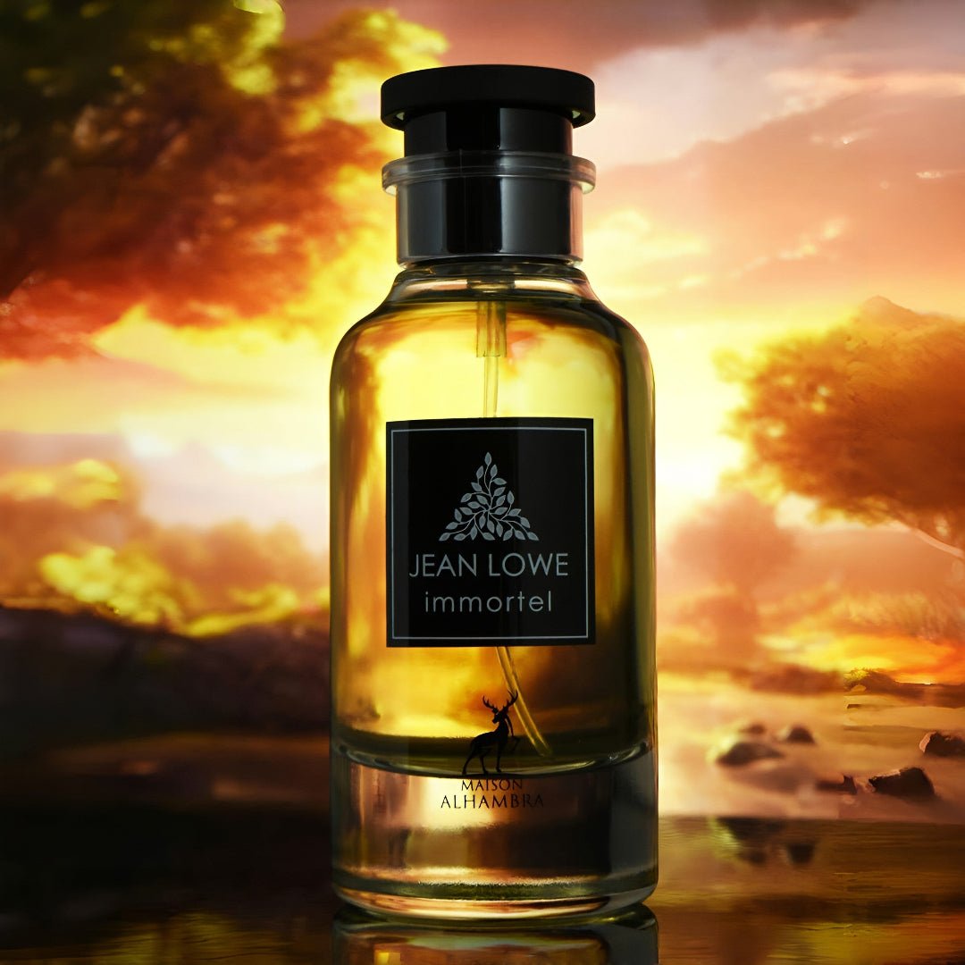 Maison Alhambra Jean Lowe Immortel — A clear glass perfume bottle with yellow liquid and a black label featuring a silver tree logo and text, set against a dramatic sunset sky with silhouetted trees and water reflections. | Maison Alhambra Jean Lowe Immortel — Botol minyak wangi kaca jernih dengan cecair kuning dan
