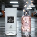 Maison Alhambra Jean Lowe Matiere — A white cylindrical box stands next to a clear glass perfume bottle filled with pink liquid and white flowers. The bottle is spraying white particles and flowers into the air. | Maison Alhambra Jean Lowe Matiere — Kotak silinder putih berdiri di sebelah botol minyak wangi kaca