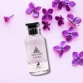 Maison Alhambra Jean Lowe Matiere — A clear glass perfume bottle with a black cap is surrounded by delicate purple lilac flowers on a light pink background. | Maison Alhambra Jean Lowe Matiere — Botol kaca jernih dengan penutup hitam dikelilingi bunga ungu lilac halus di atas latar belakang merah jambu muda.