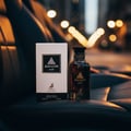 Maison Alhambra Jean Lowe Noir — A white box and a dark glass perfume bottle sit on a dark leather surface, with blurred bokeh lights in the background. | Maison Alhambra Jean Lowe Noir — Kotak putih dan botol minyak wangi kaca gelap diletakkan di atas permukaan kulit gelap, dengan lampu bokeh kabur di latar belakang.