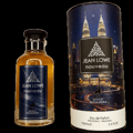 Maison Alhambra Jean Lowe Nouveau — A bottle of amber-colored perfume with a dark blue label featuring silver foliage and a deer silhouette, next to a cylindrical box with a night cityscape and twin towers | Maison Alhambra Jean Lowe Nouveau — Sebotol pewangi berwarna ambar dengan label biru gelap menampilkan siluet