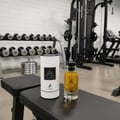 Maison Alhambra Jean Lowe Nouveau — A perfume bottle and its cylindrical packaging sit on a black gym bench, with dumbbells and gym equipment in the background. | Botol minyak wangi dan pembungkus silindernya terletak di atas bangku gim hitam, dengan dumbel dan peralatan gim di latar belakang.