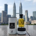 Maison Alhambra Jean Lowe Nouveau — A bottle of Jean Lowe Nouveau perfume and its cylindrical packaging stand on a stone surface with the Petronas Twin Towers in the background. | Maison Alhambra Jean Lowe Nouveau — Sebotol minyak wangi Jean Lowe Nouveau dan pembungkus silindernya berdiri di atas permukaan batu dengan