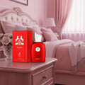Maison Alhambra Kalos by Maison Alhambra Woman 100ml — Red perfume bottle and box on a wooden nightstand next to a vase of pink roses, with a blurred pink bedroom in the background. | Maison Alhambra Kalos by Maison Alhambra Woman 100ml — Botol dan kotak pewangi merah di atas meja sisi kayu di sebelah pasu bunga mawar