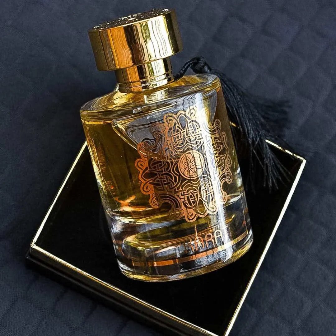 Maison Alhambra Karat by Maison Alhambra 100ml EDP — A clear glass perfume bottle with gold ornate detailing and a gold cap, resting on a black and gold square box, with a black tassel hanging from the neck. | Maison Alhambra Karat by Maison Alhambra 100ml EDP — Botol kaca jernih dengan perincian emas berornamen dan